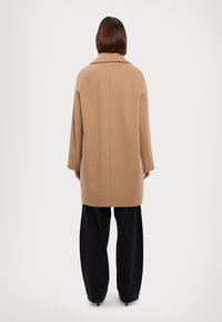 Abrigo de lana beige con un collar ancho, mangas largas y corte recto, combinado con pantalones negros holgados, visto desde la parte trasera.