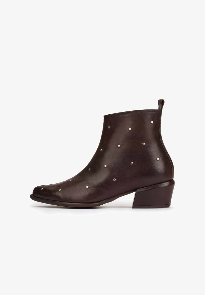 Bota de cuero marrón hasta el tobillo con punta afilada y tacón bajo cuadrado, adornada con pequeñas tachuelas metálicas espaciadas de manera uniforme por toda la superficie.