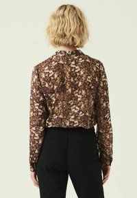 Blusa a maniche lunghe floreale in marrone scuro con motivi viola, crema e giallo; colletto con bottoni e vestibilità comoda, abbinata a pantaloni neri.
