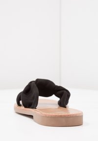 Black slip-on sandal with knotted fabric strap, tan leather insole, and flat sole. Smooth texture with a simple design and minimal accents.