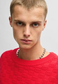 Maglione rosso lavorato a maglia con un motivo a scacchi testurizzato, colletto a girocollo a coste e collana con perline argentate. Focus sulla lavorazione a maglia e sul colore vivace.