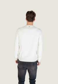 Witte geribbelde trui met lange mouwen, ronde halslijn en subtiele branding. Gecombineerd met donkergrijze slim-fit jeans.