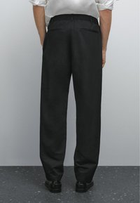 Una persona sta in piedi con le spalle rivolte verso la fotocamera, indossando una camicia bianca e pantaloni neri, su una superficie scura e testurizzata.