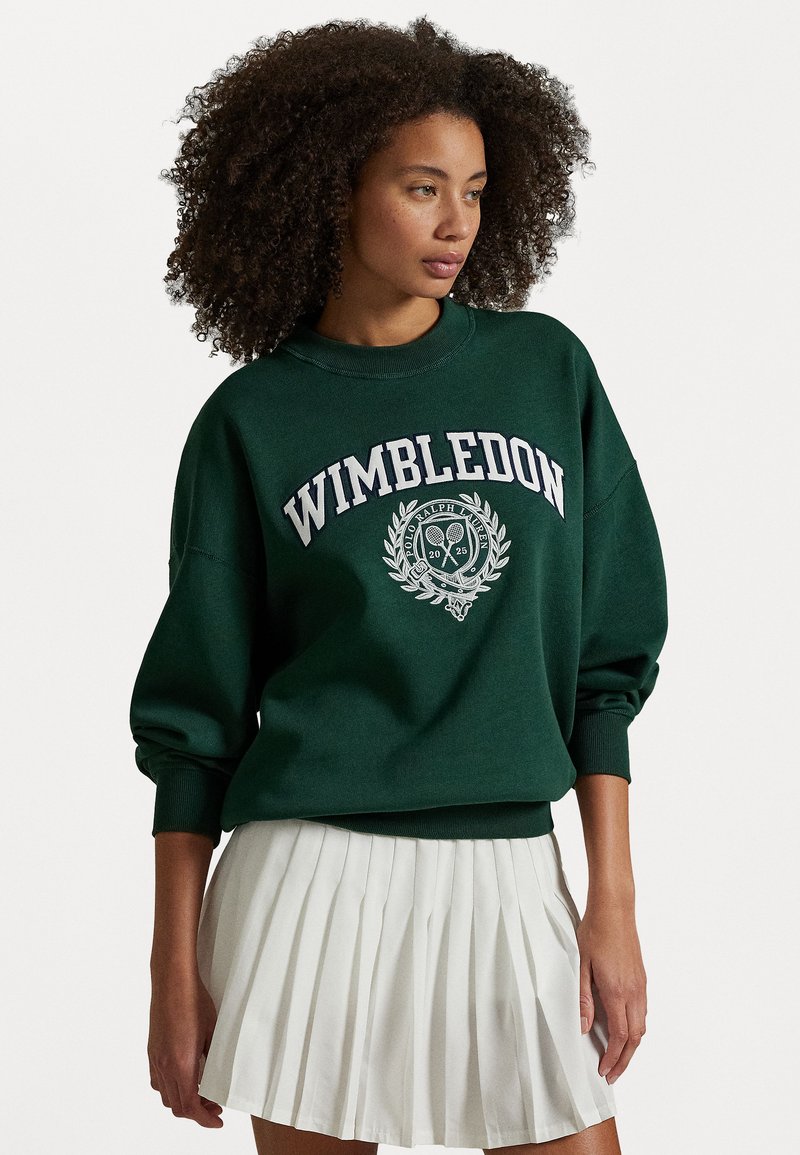Grønn hettegenser med "Wimbledon" og logo i hvit og sølv brodering; har rund hals og vide ermer. Kombineres med et hvitt plisserte skjørt.