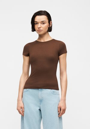 Jeune femme aux cheveux courts et foncés portant un haut marron ajusté à manches courtes et un jean bleu clair à jambes larges, debout devant un fond uni.