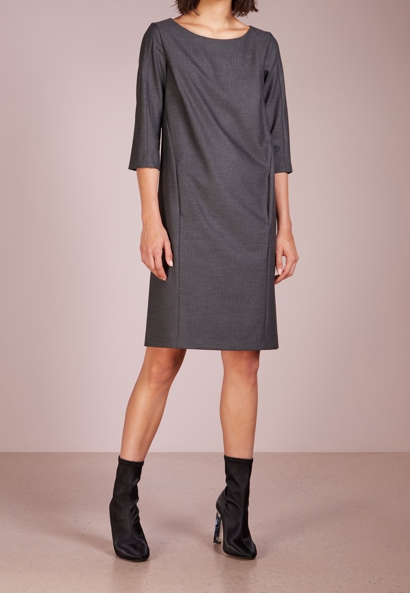 Femme portant une robe gris foncé longueur genou avec des manches trois-quarts et des bottines noires à talons hauts, debout sur un sol clair.