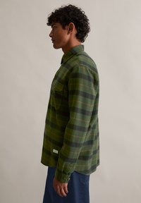 Πράσινο και μαύρο καρώ πουκάμισο από φλάνελ με πορτοκαλί λεπτομέρειες, κουμπωτές μανσέτες, μία τσέπη στο στήθος και ευθεία στρίφωμα. Φορεμένο με μπλε τζιν.