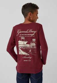Langärmliges maroon T-Shirt mit einem weißen Grafikdruck auf der Rückseite, der ein Stadion und Textdetails zu einem Spieltagsereignis zeigt.