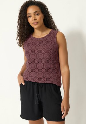 Femme aux cheveux bouclés portant un haut en crochet bordeaux sans manches et un short noir à cordon, debout devant un fond clair.