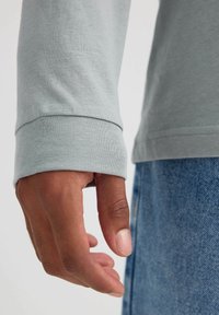 Camisa de manga larga gris claro con puños acanalados, combinada con jeans de mezclilla azul. Primer plano de una mano reposando contra la manga de la camisa.