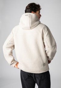 Witte fleece hoodie met een getextureerde, pluizige oppervlakte, voorzien van een capuchon en een klein merklabel dicht bij de onderste zoom. Donkere jeans worden eronder gedragen.