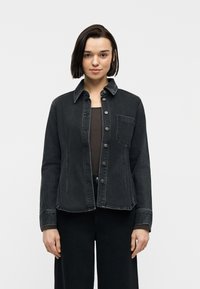 Νεαρή γυναίκα με κοντά σκούρα μαλλιά που φοράει ένα μαύρο τζιν μπουφάν με κουμπιά πάνω από σκούρο τοπ και μαύρο παντελόνι, στέκεται μπροστά σε φωτεινό μονόχρωμο φόντο.