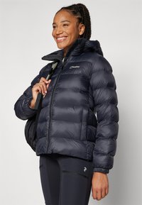 Marineblaue Pufferjacke mit hohem Kragen und Reißverschluss, mit horizontalen Steppnähten und einem Logo auf der Brust. Getragen über schwarzen Leggings.