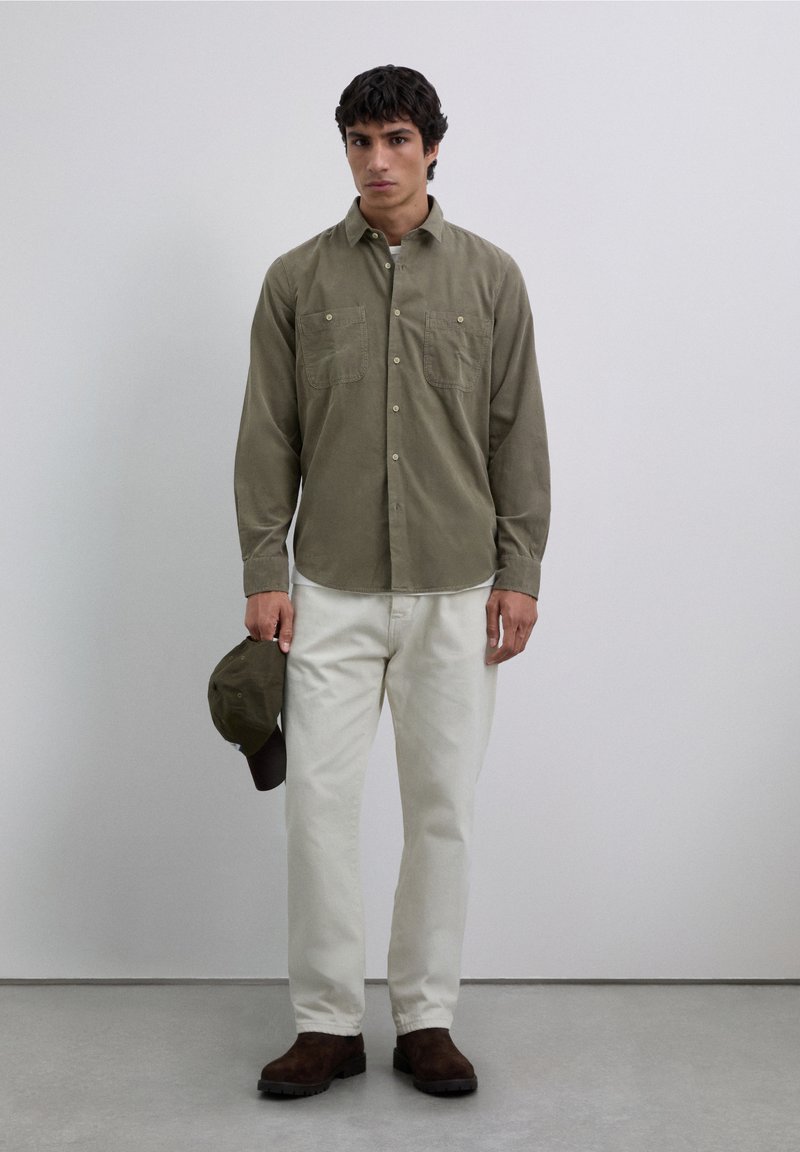 Chemise boutonnée vert olive avec deux poches poitrine, associée à un pantalon blanc et des chaussures marron, tenant une casquette assortie. Fond neutre.