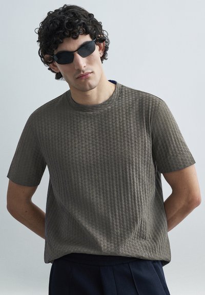 Kortærmet, tekstureret grå T-shirt med et subtilt geometrisk mønster, rund hals, båret med sorte solbriller, sat op mod en lys baggrund.