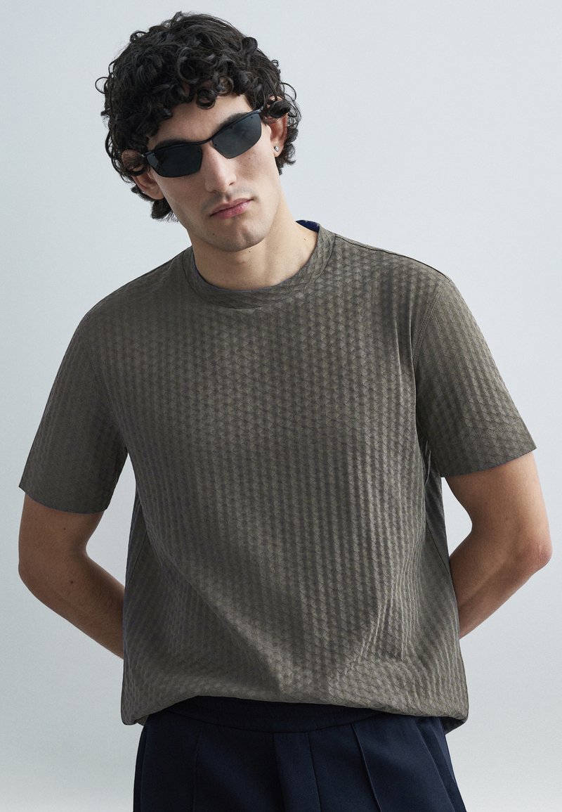 Γκρι κοντομάνικο T-shirt με υφή και διακριτικό γεωμετρικό σχέδιο, στρογγυλή λαιμόκοψη, φορεμένο με μαύρα γυαλιά ηλίου, τοποθετημένο σε ανοιχτό φόντο.