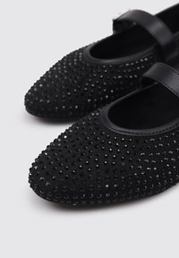 Black ballet flats with a pointed toe, featuring a textured surface embellished with small black rhinestones and a black leather strap.