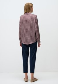 Langärmliges Hemd aus gemustertem Stoff mit roten und blauen geometrischen Formen. Kombiniert mit taillierten, marineblauen Hosen und beigen Slippern.