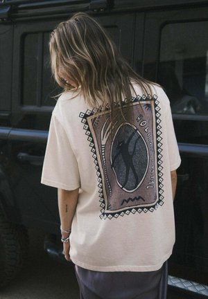 Woman with long hair wearing a beige shirt with a graphic design and text "Holiday Cocktail" on the back, standing beside a black vehicle.