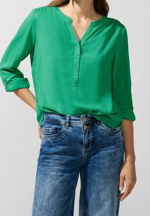 Blusa verde a maniche lunghe con scollo a V e dettagli a bottoni, abbinata a jeans in denim blu chiaro, morbidi e dal lavaggio leggero.