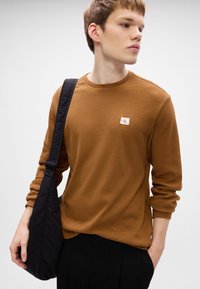 Καφέ πουκάμισο με σχέδιο waffle, μακρυμάνικο, με στρογγυλή λαιμόκοψη και μικρή λογοτύπη παράθεση, συνδυασμένο με μια μαύρη τσάντα χιαστί. Μαύρα παντελόνια ολοκληρώνουν την εμφάνιση.