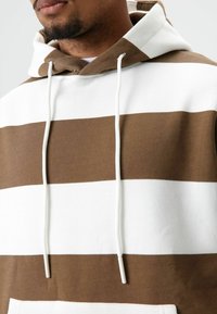 Bruine en witte gestreepte hoodie met een capuchon met trekkoord. Heeft een zachte textuur en een ontspannen pasvorm. Eenvoudig ontwerp zonder extra accenten.