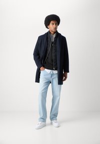 Abrigo navy sobre un chaleco puffer negro, combinado con una camisa blanca, jeans de color azul claro y zapatillas blancas. Texturas suaves y diseño en capas.