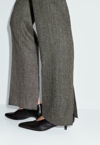 Pantaloni grigi a gamba larga con spacco laterale indossati con scarpe nere a punta con tacco kitten su uno sfondo bianco.