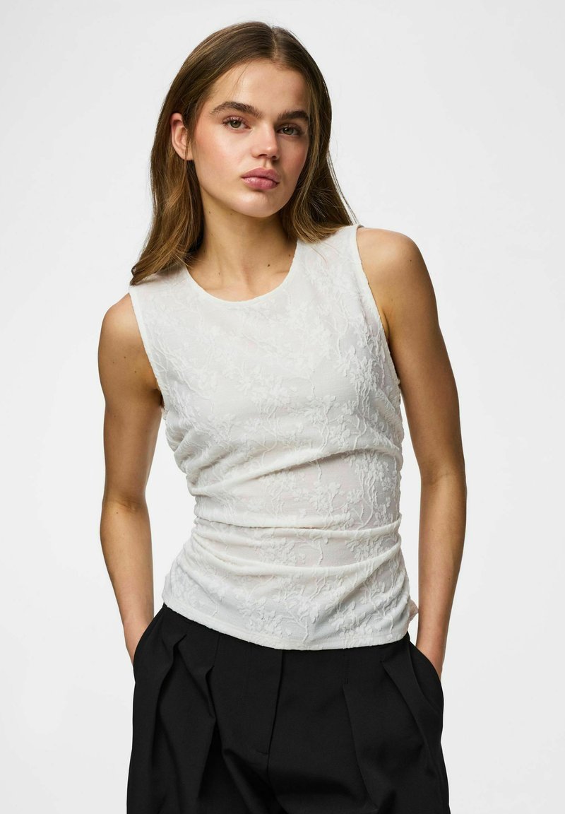 Jeune femme aux longs cheveux portant un haut blanc texturé sans manches et un pantalon noir plissé, debout les mains dans les poches.