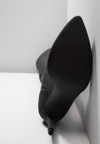 Botte noire à talon haut et bout pointu reposant sur une surface blanche, semelle tournée vers l'avant, contre un mur blanc en angle.