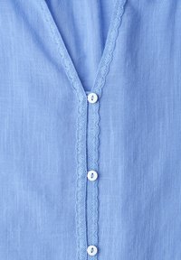 Tissu bleu clair avec une texture verticale, bordure en dentelle festonnée le long de l'ouverture frontale, et trois petits boutons blancs en rangée verticale.