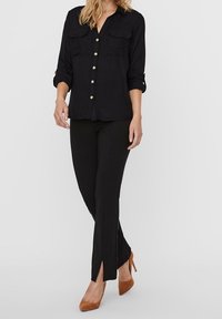 Camicia nera a bottone con due tasche frontali, abbinata a pantaloni neri aderenti con spacchetti laterali e scarpe con tacco alte marroni.