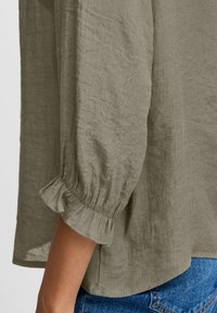 Blusa khaki a maniche lunghe in tessuto testurizzato, con polsini arricciati e una vestibilità morbida, abbinata a jeans blu.