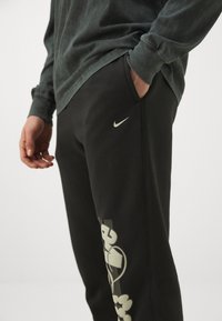 Fekete melegítőnadrág, szürke Nike logóval. Az oldalon egy merész, absztrakt minta található. Sötétzöld, hosszú ujjú pólóval kombinálva.