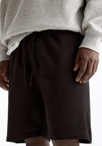 Uomo in pantaloni corti neri con cordino e felpa grigia chiara, in piedi con le mani rilassate ai lati.