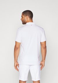 Vit kortärmad polo skjorta med krage, med en liten röd logotyp nära nacken, visad på en manlig modell från baksidan.