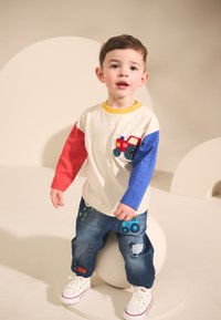 Camicia a maniche corte con maniche rosse e blu, decorata con un motivo di trattore. Abbinata a jeans blu con ricami di foglie e trattori. Scarpe da ginnastica bianche.