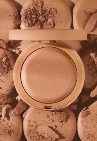 Kompaktowy bronzer o gładkiej, błyszczącej powierzchni w jasnym odcieniu opalenizny, otoczony połamanymi, matowymi, okrągłymi puderami w podobnych tonacjach.