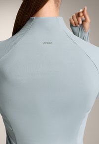 Svijetloplava atletska majica izrađena od teksturirane tkanine, s visokim ovratnikom, rebrastim detaljima i malim logom na leđima.