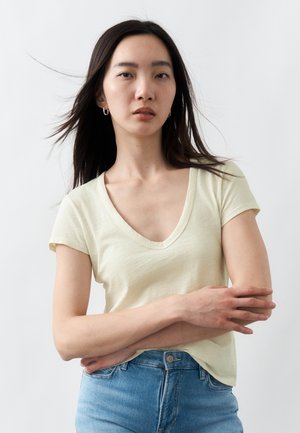 Νεαρή γυναίκα με μακριά σκούρα μαλλιά, που φοράει ανοιχτό žλεν T-shirt με V λαιμόκοψη και μπλε τζιν, με σταυρωμένα χέρια, μπροστά σε ένα απλό ανοιχτόχρωμο φόντο.