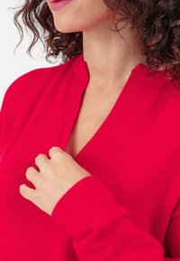 Pull-over tricoté rouge avec un col en V et des manches longues. Le tissu semble doux avec une légère texture, offrant une coupe décontractée et confortable.