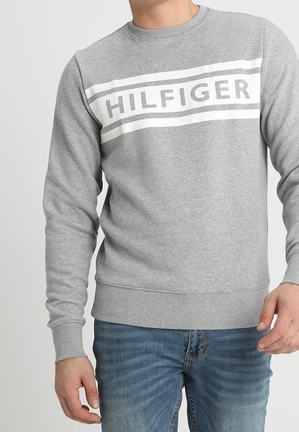 Uomo vestito con una felpa grigia con "HILFIGER" in lettere bianche in grassetto sul petto e jeans blu.