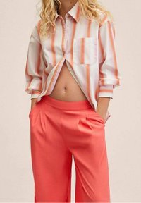 Gestreept overhemd in wit, roze en oranje met een borstzak, gecombineerd met koral gekleurde plooibroek met wijde pijpen.