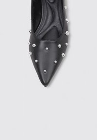 Zapato negro de punta afilada hecho de cuero suave, con tachuelas plateadas dispuestas decorativamente en la parte superior. Plantilla suave y acolchada.