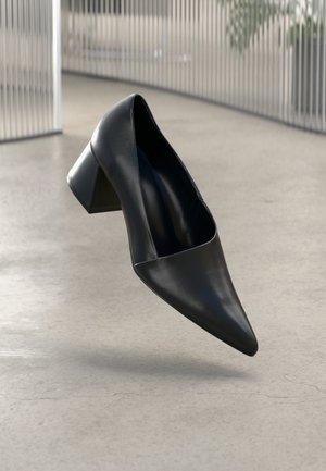 Scarpa nera in pelle con tacco, punta affusolata e tacco spesso. Presenta un design elegante e una texture opaca, sospesa sopra una superficie liscia.