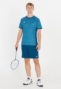 Ung man som ler, står med ett badmintonracket, iförd en blåmönstrad sporttröja, matchande shorts, vita strumpor och sneakers.