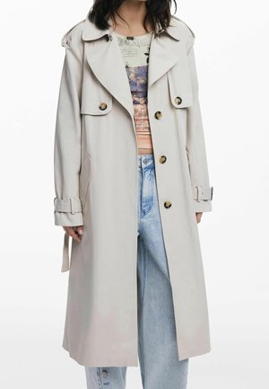 Femme portant un long trench-coat beige clair avec de grands revers, un devant boutonné et des poignets ceinturés, sur un haut à motif et un jean bleu clair.