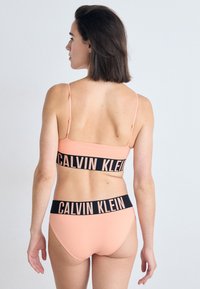 Calvin Klein sportbeha en bijpassende slip in zacht perzik met zwarte logobanden. De beha heeft dunne banden; de slip heeft een laag uitgesneden model.