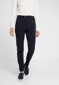 Pantalons stretch navy avec une texture lisse, coupe slim, deux poches zippées à l'avant et une fermeture à boutons. Associés à un léger pull et des chaussures décontractées.