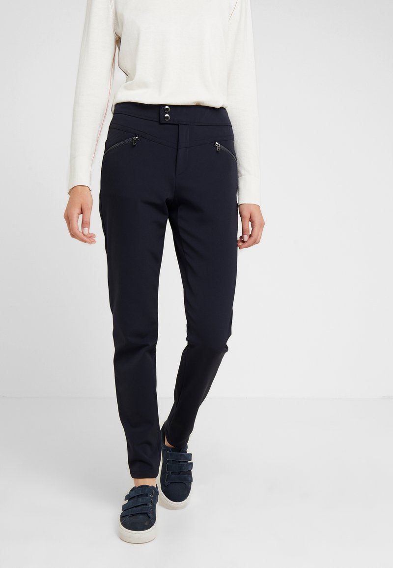 Pantalons stretch navy avec une texture lisse, coupe slim, deux poches zippées à l'avant et une fermeture à boutons. Associés à un léger pull et des chaussures décontractées.